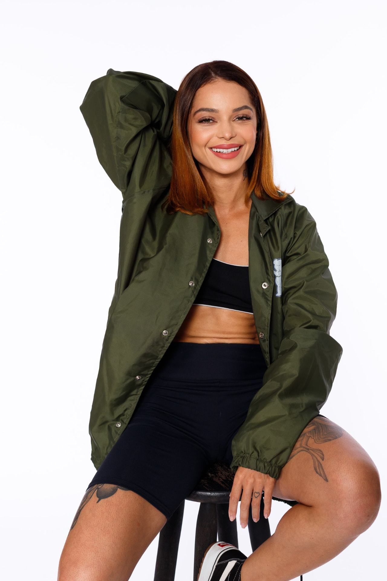 Bárbara Macedo sorrindo de jaqueta verde e look esportivo, em estúdio